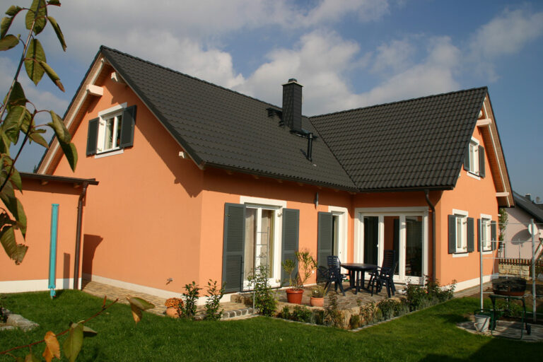 Ein modernes, pfirsichfarbenes Haus mit einem dunkelgrauen Ziegeldach, weiß gerahmten Fenstern mit Fensterläden, einer Terrasse mit Gartenmöbeln und einem grünen Rasen unter einem teilweise bewölkten Himmel.