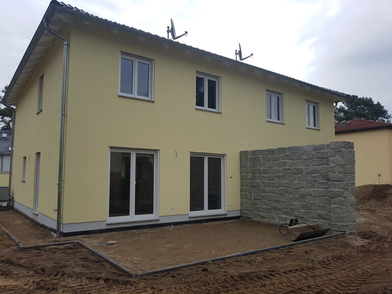 Ein neu erbautes gelbes Doppelhaus mit weiß gerahmten Fenstern und Türen, einer Steinmauer auf einer Seite und einem unvollendeten, unbefestigten Hof im Vordergrund.